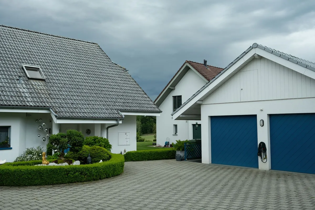 Weiß verputztes Einfamilienhaus mit Doppelgarage und blauen Toren, davor eine breite gepflasterte Einfahrt mit Heckenrand