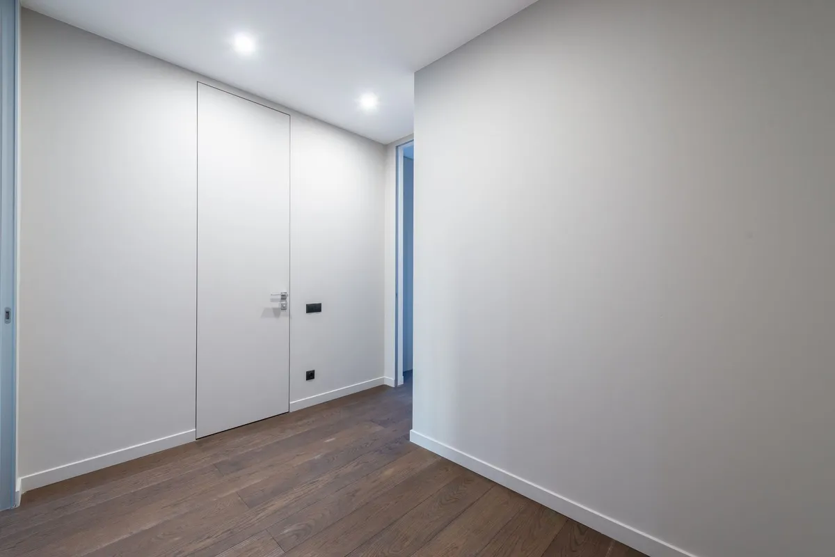 Leerer Wohnungsflur mit weißen Wänden, dunklem Holzdielenboden, flächenbündiger weißer Tür und zwei Deckenspots