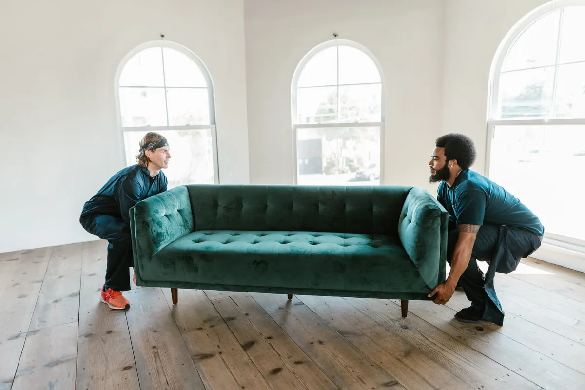 Zwei Umzugshelfer setzen gemeinsam ein großes grünes Sofa in einem leeren Raum mit alten Holzdielen ab