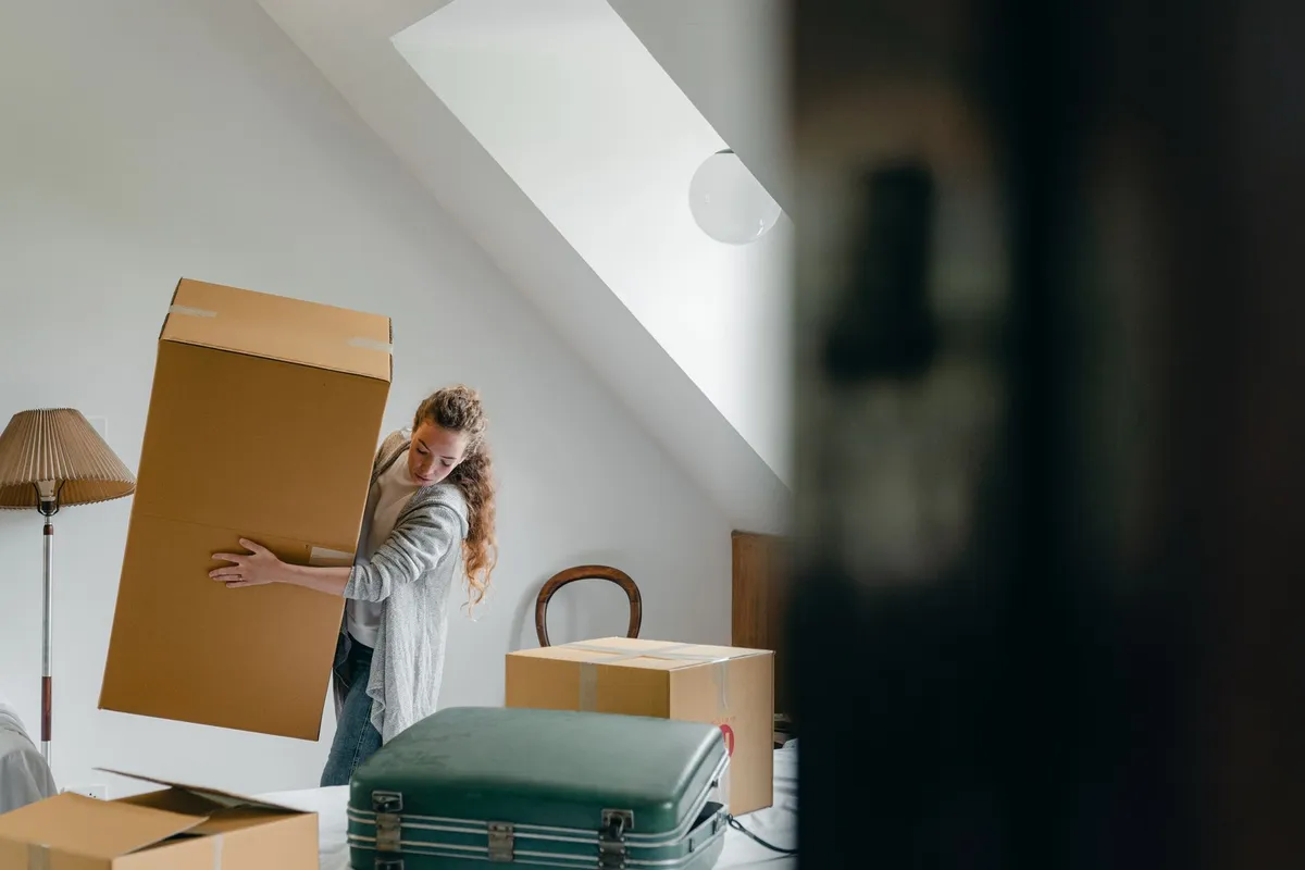 Frau trägt einen großen Umzugskarton durch ein altmodisch eingerichtetes Dachzimmer mit Holzstuhl, Stehlampe und altem Koffer