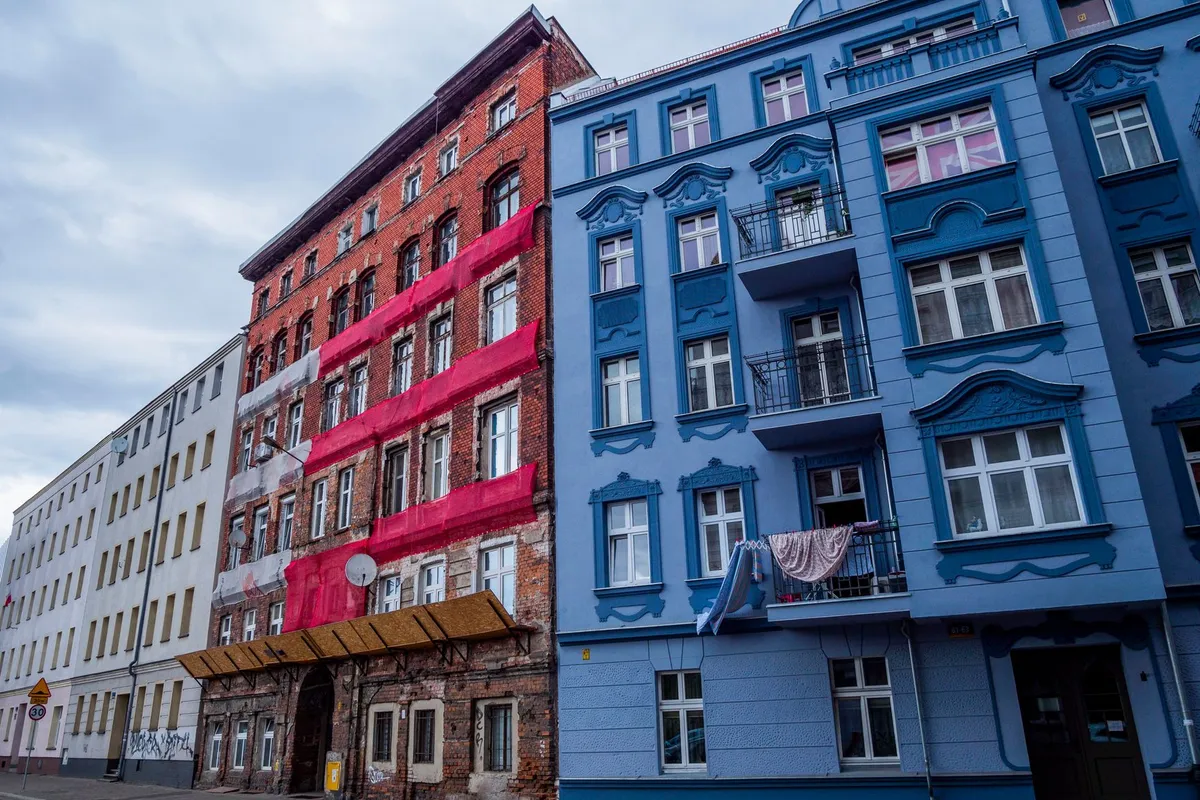 Dichte Straßenzeile mit fünfgeschossigen Gründerzeithäusern, links roter Backsteinbau, rechts blaue Stuckfassade mit kleinen Balkonen